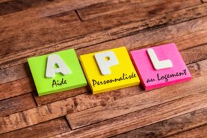 Trois post-it colorés alignés formant « APL » avec l’inscription « Aide Personnalisée au Logement » sur fond en bois.