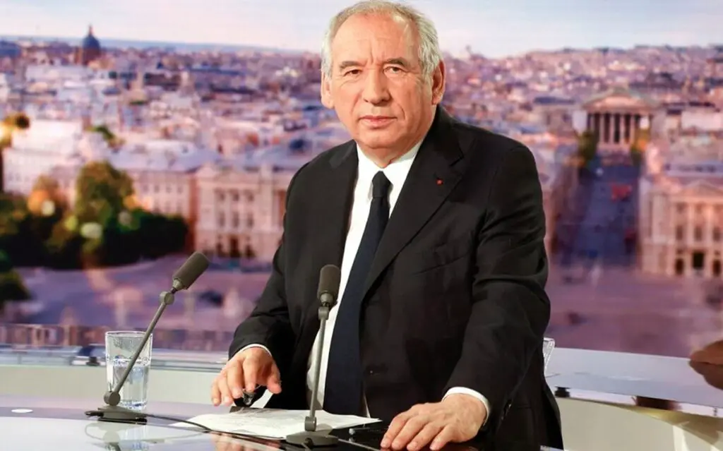 Bayrou assis sur un plateau télé, devant deux micros et des feuilles, avec en arrière-plan une vue floue de Paris.
