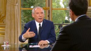 François Bayrou en costume bleu, de face, parlant et gesticulant lors d’une interview télévisée ; l’intervieweur est vu de dos, grande fenêtre et jardin en arrière-plan.