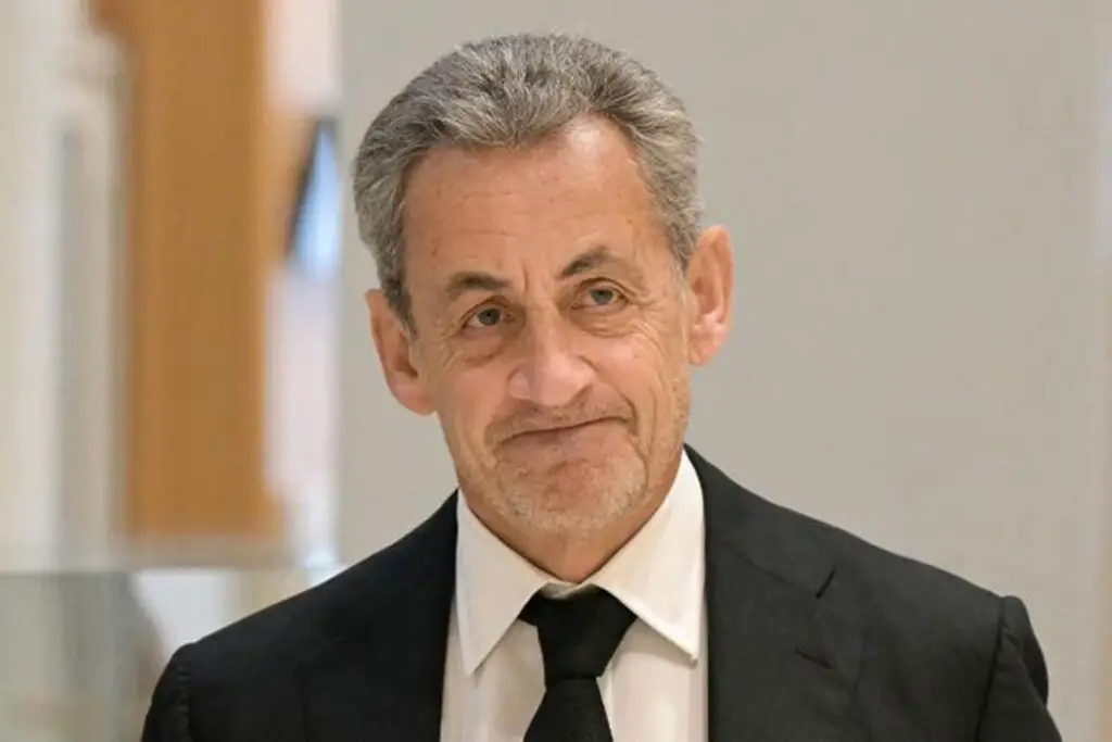 Sarkozy vêtu d'un costume sombre et cravate noire, visage de trois-quarts, expression sérieuse, en intérieur.