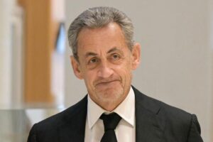 Sarkozy vêtu d'un costume sombre et cravate noire, visage de trois-quarts, expression sérieuse, en intérieur.