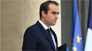 Sébastien Lecornu en costume sombre, de profil, marchant devant un bâtiment officiel ; drapeaux français et européen flous à l’arrière-plan.