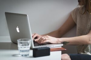Personne tapant sur un ordinateur portable posé sur un bureau, avec un verre d’eau au premier plan et un petit haut-parleur ; ambiance intérieure, cadrage sur les mains.