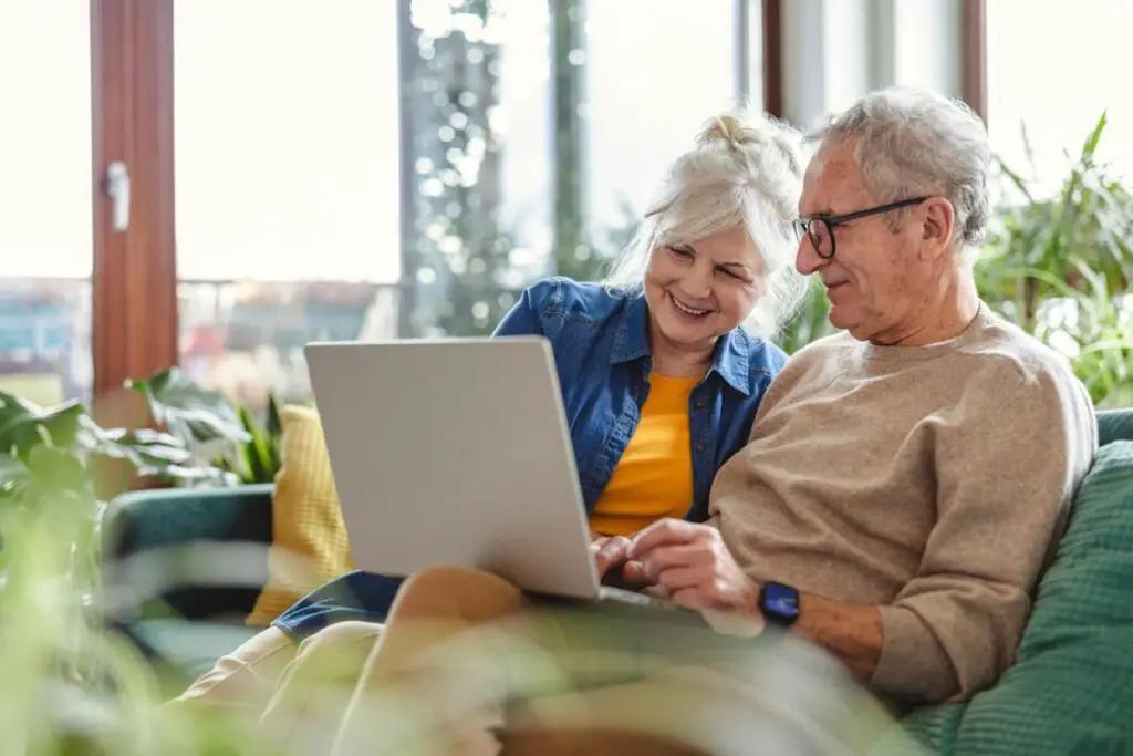 Couple de seniors souriants assis sur un canapé, consultant un ordinateur portable dans un salon lumineux avec des plantes.