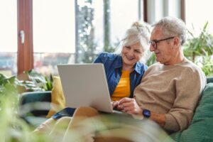 Couple de seniors souriants assis sur un canapé, consultant un ordinateur portable dans un salon lumineux avec des plantes.