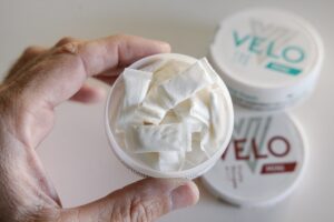Gros plan d’une main tenant une boîte ouverte de sachets de nicotine blancs, avec des boîtes « VELO » floutées à l’arrière-plan.